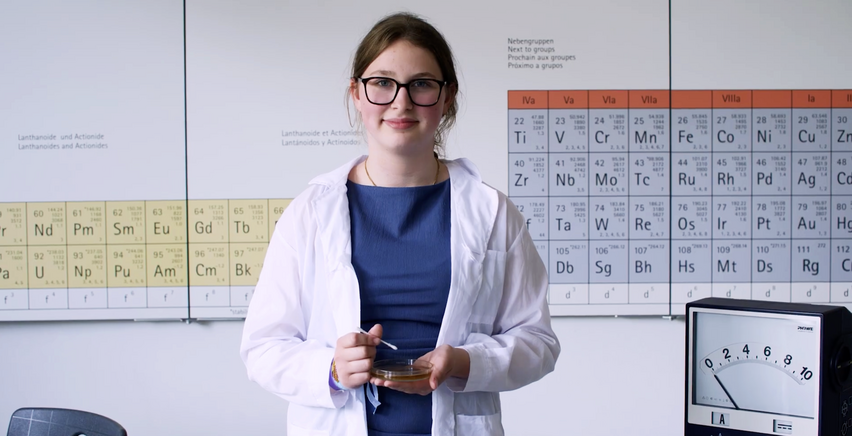 Eine Schülerin im Laborkittel steht vor einer Wand mit einem Periodensystem