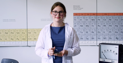 Eine Schülerin im Laborkittel steht vor einer Wand mit einem Periodensystem