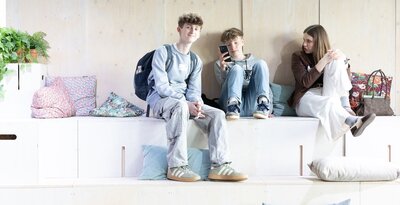 Drei Schüler:innen sitzen zusammen auf einer großen Holztreppe mit Kissen